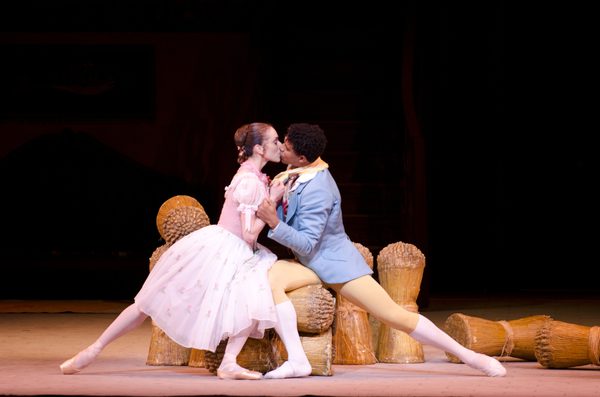 Two people sit on a pile of stooks of corn. The figure on the left is Lise, she wears a pale pink dress. The figure on the right is Colas, he wears a blue jacked and yellow tights. They are kissing.