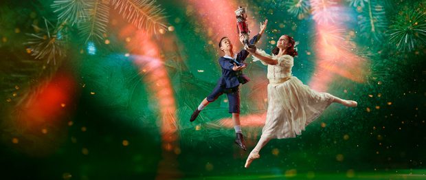 There are two dancers, slightly to the right of centre. The background is dark green with a large, brighter spotlight that doesn't reach the bottom corners of the image. In the background are giant pine needles, a scattering of small lighter areas, and curved beams of red light, that look paler when they are within the spotlight. The dancer in the foreground has shoulder length brown hair tied back with a white ribbon. She is representing the character of Clara. She is facing towards the left of the picture. She wears a white dress and a pair of off-white ballet pointe shoes. She is in mid leap with her left leg stretched out behind her. Her right leg is pointing towards the floor. She's holding up in front of her with both hands a Nutcracker doll, painted to look like a soldier in a red jacket and white trousers. The dancer behind Clara is representing the character of Fritz, Clara's younger brother. He has short dark hair and is wearing a dark blue sailor suit with britches to just below his knee. On his feet are white socks and soft black ballet shoes. He is facing Clara and his arms are raised as if trying to steal the Nutcracker doll from her. Photo copyright Manvir Rai.