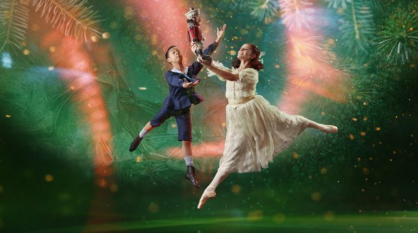 There are two dancers, slightly to the right of centre. The background is dark green with a large, brighter spotlight that doesn't reach the bottom corners of the image. In the background are giant pine needles, a scattering of small lighter areas, and curved beams of red light, that look paler when they are within the spotlight. The dancer in the foreground has shoulder length brown hair tied back with a white ribbon. She is representing the character of Clara. She is facing towards the left of the picture. She wears a white dress and a pair of off-white ballet pointe shoes. She is in mid leap with her left leg stretched out behind her. Her right leg is pointing towards the floor. She's holding up in front of her with both hands a Nutcracker doll, painted to look like a soldier in a red jacket and white trousers. The dancer behind Clara is representing the character of Fritz, Clara's younger brother. He has short dark hair and is wearing a dark blue sailor suit with britches to just below his knee. On his feet are white socks and soft black ballet shoes. He is facing Clara and his arms are raised as if trying to steal the Nutcracker doll from her. Photo copyright Manvir Rai.