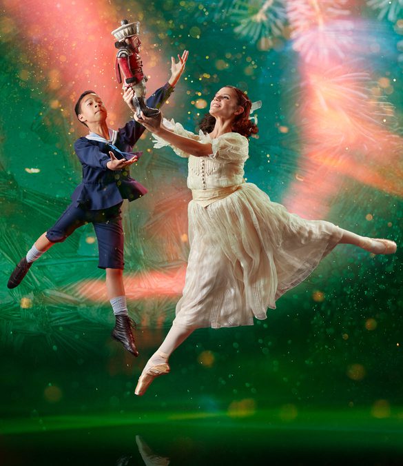 There are two dancers, slightly to the right of centre. The background is dark green with a large, brighter spotlight that doesn't reach the bottom corners of the image. In the background are giant pine needles, a scattering of small lighter areas, and curved beams of red light, that look paler when they are within the spotlight. The dancer in the foreground has shoulder length brown hair tied back with a white ribbon. She is representing the character of Clara. She is facing towards the left of the picture. She wears a white dress and a pair of off-white ballet pointe shoes. She is in mid leap with her left leg stretched out behind her. Her right leg is pointing towards the floor. She's holding up in front of her with both hands a Nutcracker doll, painted to look like a soldier in a red jacket and white trousers. The dancer behind Clara is representing the character of Fritz, Clara's younger brother. He has short dark hair and is wearing a dark blue sailor suit with britches to just below his knee. On his feet are white socks and soft black ballet shoes. He is facing Clara and his arms are raised as if trying to steal the Nutcracker doll from her. Photo copyright Manvir Rai.