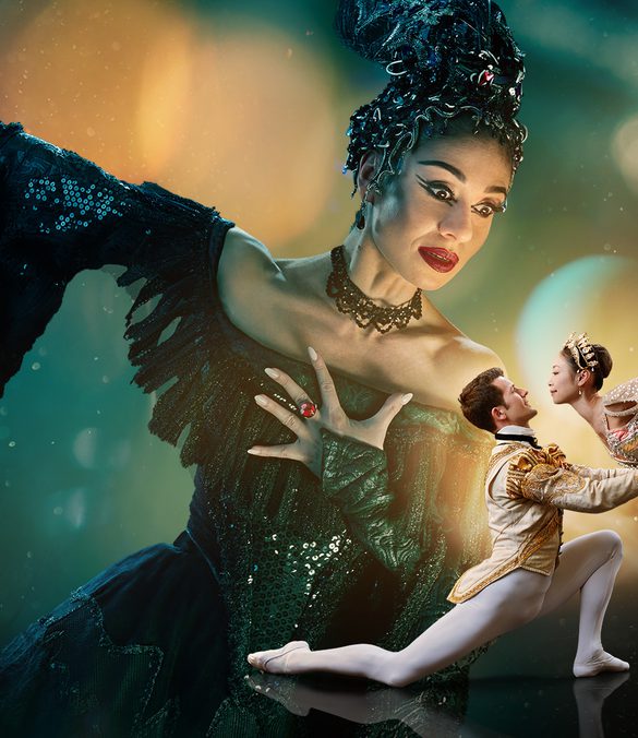 Two dancers are in the foreground looking at each other with a much larger figure in the background towering over them. The background is a blended blue and green at the top of the image. The image fades to black at the bottom. Surrounding the two dancers in the foreground is a glowing gold light with spots and streams of light surrounding the centre of the image. This gold is reflected onto the towering figure’s face. The two dancers in the foreground are towards the right of the image and are playing the characters of the Prince and Sleeping Beauty. The dancer playing the Prince has brown, short hair, is wearing white, fitted tights, white ballet flats and a cream jacket with gold detailing. He is on the left of the couple, kneeling one knee, looking up at Sleeping Beauty, with arms extended, holding her waist. The dancer playing Sleeping Beauty has dark hair in a bun, is wearing a white tutu with silver detailing, a sequinned top, sparkling tiara and is on pointe on her right leg, with her left leg extended pointing directly upwards. The figure in the background is playing Carabosse, she is positioned to the right of the foreground dancers, and is looking down at them. The dancer playing Carabosse has black hair with a silver headpiece, is wearing a gothic black dress with sequinned detailing and has black eye-makeup and red lipstick. Carabosse has her left arm clutching at her chest, a ring with large, red gemstone is on her middle finger, her right arm is extended towards the upper left and a dramatic black sleeve drapes towards the floor.