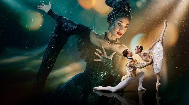 Two dancers are in the foreground looking at each other with a much larger figure in the background towering over them. The background is a blended blue and green at the top of the image. The image fades to black at the bottom. Surrounding the two dancers in the foreground is a glowing gold light with spots and streams of light surrounding the centre of the image. This gold is reflected onto the towering figure’s face. The two dancers in the foreground are towards the right of the image and are playing the characters of the Prince and Sleeping Beauty. The dancer playing the Prince has brown, short hair, is wearing white, fitted tights, white ballet flats and a cream jacket with gold detailing. He is on the left of the couple, kneeling one knee, looking up at Sleeping Beauty, with arms extended, holding her waist. The dancer playing Sleeping Beauty has dark hair in a bun, is wearing a white tutu with silver detailing, a sequinned top, sparkling tiara and is on pointe on her right leg, with her left leg extended pointing directly upwards. The figure in the background is playing Carabosse, she is positioned to the right of the foreground dancers, and is looking down at them. The dancer playing Carabosse has black hair with a silver headpiece, is wearing a gothic black dress with sequinned detailing and has black eye-makeup and red lipstick. Carabosse has her left arm clutching at her chest, a ring with large, red gemstone is on her middle finger, her right arm is extended towards the upper left and a dramatic black sleeve drapes towards the floor.