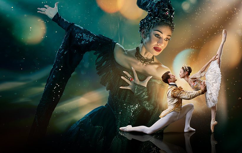 Two dancers are in the foreground looking at each other with a much larger figure in the background towering over them. The background is a blended blue and green at the top of the image. The image fades to black at the bottom. Surrounding the two dancers in the foreground is a glowing gold light with spots and streams of light surrounding the centre of the image. This gold is reflected onto the towering figure’s face. The two dancers in the foreground are towards the right of the image and are playing the characters of the Prince and Sleeping Beauty. The dancer playing the Prince has brown, short hair, is wearing white, fitted tights, white ballet flats and a cream jacket with gold detailing. He is on the left of the couple, kneeling one knee, looking up at Sleeping Beauty, with arms extended, holding her waist. The dancer playing Sleeping Beauty has dark hair in a bun, is wearing a white tutu with silver detailing, a sequinned top, sparkling tiara and is on pointe on her right leg, with her left leg extended pointing directly upwards. The figure in the background is playing Carabosse, she is positioned to the right of the foreground dancers, and is looking down at them. The dancer playing Carabosse has black hair with a silver headpiece, is wearing a gothic black dress with sequinned detailing and has black eye-makeup and red lipstick. Carabosse has her left arm clutching at her chest, a ring with large, red gemstone is on her middle finger, her right arm is extended towards the upper left and a dramatic black sleeve drapes towards the floor.