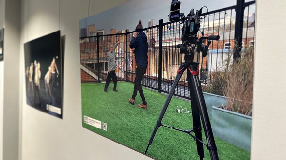 Henry McNab of the Freefall @ 21 exhibition at Birmingham Hippodrome. Dancers of the Freefall Dance Company are pictured in two different pictures hung against a grey wall. In the focal image, one of the dancers is amongst 5 white chairs in a green field surrounded with mature trees at the edges. The dancer is facing the camera with arms are spread outwards, and they are looking to the right of the image. There is more information in the bottom right corner. The image to the left of the focal image is out of focus, but pictures a group of the Freefall dancers wearing black in a dance studio. Credit: Henry McNab