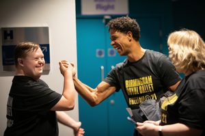 A member of Freefall shaking Carlos Acosta’s hand backstage at Birmingham Hippodrome. The member of Freefall is on the left, shaking Carlos’ right hand. The Freefaller smiling whilst standing side-on to the camera and has short brown hair, a hearing aid in the right ear and is wearing a black t-shirt with ‘Birmingham Royal Ballet, Freefall Dance’ written in white on the back. Carlos smiling whilst shaking the Freefaller’s right hand and has the arm bent at the elbow. Carlos has short, dark, curly hair and is wearing a black t-shirt with ‘Birmingham Royal Ballet, Freefall @ 21’ written in yellow across the front. Carlos’ grey, checked coat is draped over the left arm. To the right of Carlos is Claire, who has shoulder-length blonde hair and is wearing a similar t-shirt to Carlos, holding a phone with a dark blue case in both hands. In the background there is a blue set of double doors to the right and a white wall with a silver sign to the left. Credit: John Palmer.