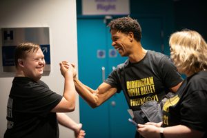 A member of Freefall shaking Carlos Acosta’s hand backstage at Birmingham Hippodrome. The member of Freefall is on the left, shaking Carlos’ right hand. The Freefaller smiling whilst standing side-on to the camera and has short brown hair, a hearing aid in the right ear and is wearing a black t-shirt with ‘Birmingham Royal Ballet, Freefall Dance’ written in white on the back. Carlos smiling whilst shaking the Freefaller’s right hand and has the arm bent at the elbow. Carlos has short, dark, curly hair and is wearing a black t-shirt with ‘Birmingham Royal Ballet, Freefall @ 21’ written in yellow across the front. Carlos’ grey, checked coat is draped over the left arm. To the right of Carlos is Claire, who has shoulder-length blonde hair and is wearing a similar t-shirt to Carlos, holding a phone with a dark blue case in both hands. In the background there is a blue set of double doors to the right and a white wall with a silver sign to the left. Credit: John Palmer.