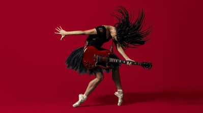 BRB Dancer Sofia Liñares is standing in the centre of the image, against a deep red background on which her shadow can be seen. Sofia Liñares is a dancer with long dark hair and is wearing a black sleeveless top, with black tutu, and pale pink pointe shoes. Sofia is standing with her left foot on pointe, her right leg is slightly bent at the knee, the top of her foot is resting on the ground. Sofia’s head is mid-motion, facing towards the ground, her hair is flying forwards with the strands of hair in an arch around her head. Sofia is holding a red and black electric guitar which is secured over her shoulder using a strap decorated with red roses and green leaves. Sofia’s right arm is facing the left as if she had just strummed the guitar, her left is holding the fret board in a chord. Image copyright Perou.