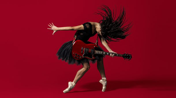 BRB Dancer Sofia Liñares is standing in the centre of the image, against a deep red background on which her shadow can be seen. Sofia Liñares is a dancer with long dark hair and is wearing a black sleeveless top, with black tutu, and pale pink pointe shoes. Sofia is standing with her left foot on pointe, her right leg is slightly bent at the knee, the top of her foot is resting on the ground. Sofia’s head is mid-motion, facing towards the ground, her hair is flying forwards with the strands of hair in an arch around her head. Sofia is holding a red and black electric guitar which is secured over her shoulder using a strap decorated with red roses and green leaves. Sofia’s right arm is facing the left as if she had just strummed the guitar, her left is holding the fret board in a chord. Image copyright Perou.