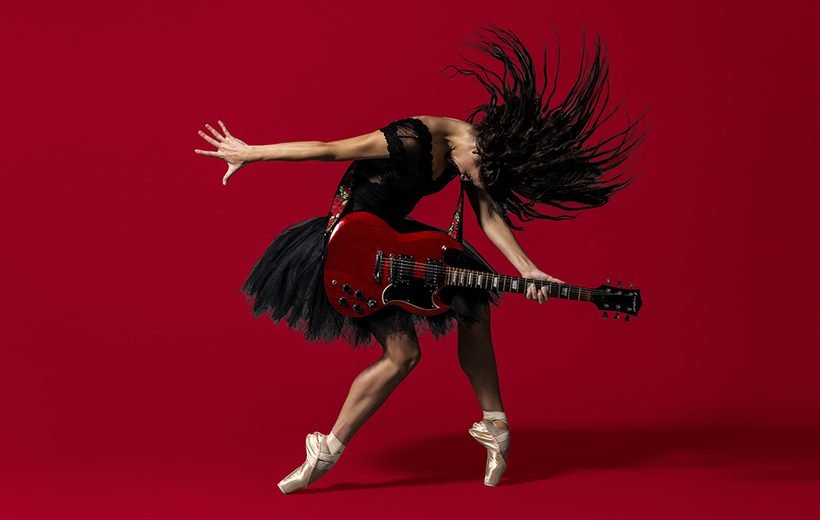 BRB Dancer Sofia Liñares is standing in the centre of the image, against a deep red background on which her shadow can be seen. Sofia Liñares is a dancer with long dark hair and is wearing a black sleeveless top, with black tutu, and pale pink pointe shoes. Sofia is standing with her left foot on pointe, her right leg is slightly bent at the knee, the top of her foot is resting on the ground. Sofia’s head is mid-motion, facing towards the ground, her hair is flying forwards with the strands of hair in an arch around her head. Sofia is holding a red and black electric guitar which is secured over her shoulder using a strap decorated with red roses and green leaves. Sofia’s right arm is facing the left as if she had just strummed the guitar, her left is holding the fret board in a chord. Image copyright Perou.