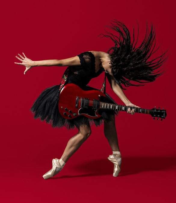 BRB Dancer Sofia Liñares is standing in the centre of the image, against a deep red background on which her shadow can be seen. Sofia Liñares is a dancer with long dark hair and is wearing a black sleeveless top, with black tutu, and pale pink pointe shoes. Sofia is standing with her left foot on pointe, her right leg is slightly bent at the knee, the top of her foot is resting on the ground. Sofia’s head is mid-motion, facing towards the ground, her hair is flying forwards with the strands of hair in an arch around her head. Sofia is holding a red and black electric guitar which is secured over her shoulder using a strap decorated with red roses and green leaves. Sofia’s right arm is facing the left as if she had just strummed the guitar, her left is holding the fret board in a chord. Image copyright Perou.