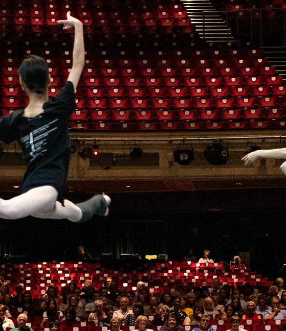 Looking from the back of the stage towards the auditorium, two dancers are leaping on the stage watched by an audience seated in the auditorium