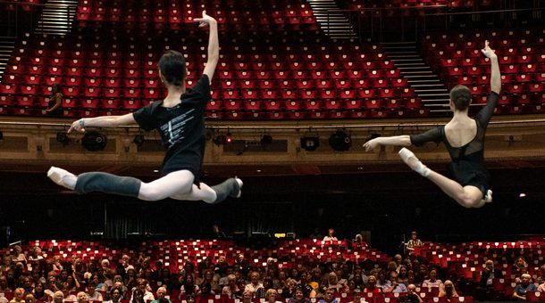 Looking from the back of the stage towards the auditorium, two dancers are leaping on the stage watched by an audience seated in the auditorium