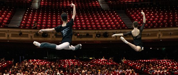 Looking from the back of the stage towards the auditorium, two dancers are leaping on the stage watched by an audience seated in the auditorium