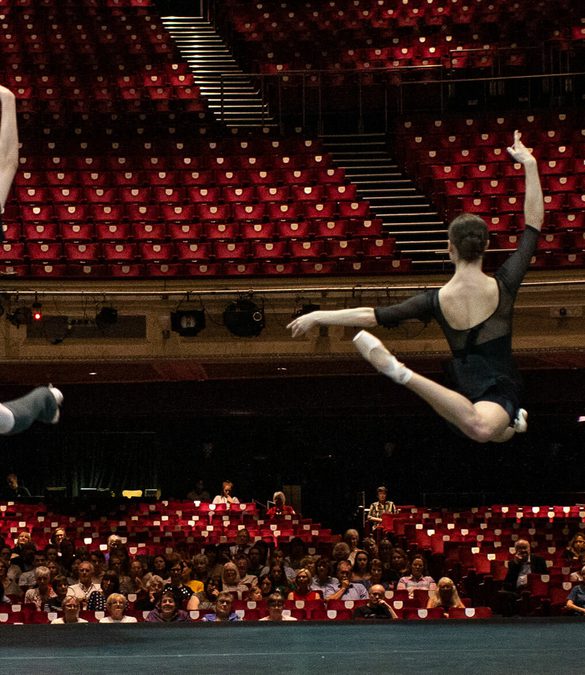 Looking from the back of the stage towards the auditorium, two dancers are leaping on the stage watched by an audience seated in the auditorium