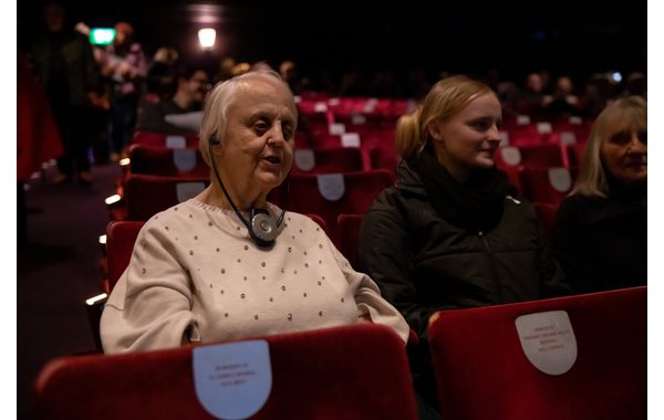 This image shows a theatre auditorium. To the left of the image in the foreground is a visually impaired person with a headset listening to the audio describer's commentary. The headset is black with discreet headphones attached to a silver button which adjusts the volume. Next to them are a few audience members.