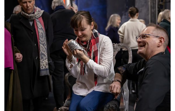 A young woman is seated in the centre of the image holding a silver slipper and touching the sole of it. The audio describer is kneeling down to the right of her smiling and looking at someone to the left of the image.