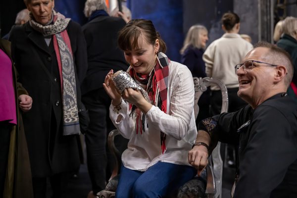 A young woman is seated in the centre of the image holding a silver slipper and touching the sole of it. The audio describer is kneeling down to the right of her smiling and looking at someone to the left of the image.