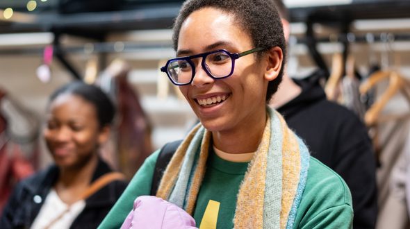 A young woman is in the centre smiling and looking to her right. She has short black hair and is wearing black glasses. She has a pink bag in her arms. There is another young woman to the left of her. Behind them is a clothes rail slightly out of focus with clothing on hangers.