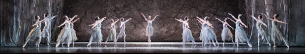 The Snow Fairy stands in the middle of the stage dressed in a kneelength white dresson point and with her arms raised up, with a dozen female and male dancers on either side in a row, facing the Snow Fairy one arm raised pointing towards her, the other lower and behind