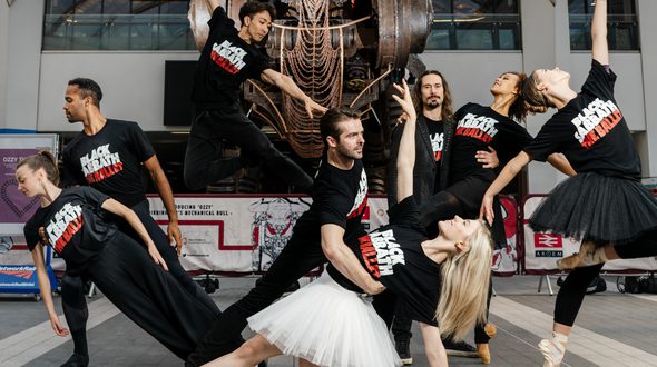 BRB Dancers pose in front of ‘Ozzy’ the Bull in Grand Central Station, Birmingham wearing t-shirt which say ‘Black Sabbath The Ballet’ on. 8 BRB dancers are posing in front of the huge metalwork bull which featured in the Birmingham Commonwealth Games opening ceremony. The dancers are in various poses from the show, some standing, jumping or kneeling on the grey tile concourse. The dancers are wearing black t-shirts with Black Sabbath (in white), The Ballet (in red) on the front. Some of the dancers are in pale pink pointe shoes and white or black tutus. ‘Ozzy’ is looming over the dancers, with enormous metal horns and mechanical eyes. In the background, the white arched ceilings of the station can be seen, below which is a series of black framed windows. Credit Kris Askey.