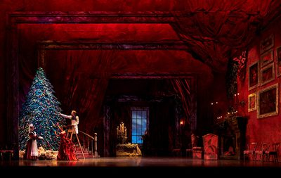 We're in the hallway of the Stahlbaums' beautiful Victorian home. To the left is a giant Christmas tree, standing next to a sweeping staircase. There are presents under the tree, and one of the servants is standing on a stepladder lighting the candles on the tree while Mrs Stahlbaum supervises and one of the maids stands by. On the other side of the room is a grand fireplace set into a wall covered with many large paintings in ornate gold frames. The wallpaper throughout the room is a rich red. In the centre to the back is a window showing a dark blue night sky.