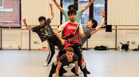 Enrique Bejarano Vidal, Gus Payne, Javier Rojas, Raúl Reinoso, Shuailun Wu in rehearsal of Black Sabbath - The Ballet. In this group photo, Gus (who has brown hair with blonde highlights and is wearing a grey top and black watch) is in a press-up position underneath Raúl’s legs (Raúl has dark, short, curly hair, dark stubble and is wearing a red t-shirt with white sleeves which reads ‘Habano’ across the chest, black tracksuit bottoms with white and red detailing and black socks). Raúl is holding a red and black electric guitar, with left hand on the fretboard. Shuailun (who has short dark hair and is wearing a grey t-shirt) is on the right of Raúl, leaning backwards with left arm arched behind the head. Javier is on the left of Raúl, (Javier has short, dark, curly hair, is wearing a dark grey t-shirt with gold lettering and faces on, black tracksuit bottoms which have a white panel on the sides. Black buttons run all the way down the leg and three black stripes are top of the white panel. Javier also has a silver chain necklace and white socks). Javier has both arms raised, the left higher than the right, a bent right knee and is leaning out towards the left of the image. Enrique is being masked by Raúl’s head, but the top of their dark short hair can be seen. Enrique has both arms raised in a v-shape and is wearing a teal blue t-shirt. In the background there are dark framed windows, between which are white panelled walls. A ballet barre runs across the rear of the room and dancer’s equipment is sat beneath it. Credit Johan Persson.