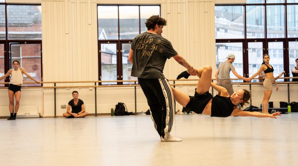 BRB Dancers Javier Rojas and Yaoqian Shang are mid-movement during rehearsals for Black Sabbath - The Ballet. Javier has short, dark, curly hair, is wearing a dark grey t-shirt with gold lettering on the back which reads ‘drop it like it’s hot,’ black tracksuit bottoms which have a white panel on the sides. Black buttons run all the way down the leg and three black stripes are top of the white panel. Javier also has a silver chain necklace and white socks on. Javier is standing almost upright and with right arm outstretched, is holding Yaoqian’s right arm, hands clasped just below Javier’s arm tattoos. Yaoqian has dark hair with blonde highlights and is wearing a black crop top, black shorts, black socks and a silver earring in the left ear. Yaoqian is being held about 10 inches off of the ground with body parallel to the grey Lino floor. Yaoqian’s left arm is outstretched towards the right of the image and the right arm is underneath the right leg, which is bent at the knee. The left leg is hidden behind Javier’s torso. In the background, three large, dark-framed windows light the studio. White panelled walls sit in the middle of the windows, and a ballet barre runs across the back wall. Five dancers watch Javier and Yaoqian in rehearsal. Credit Johan Persson.