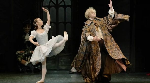 Two dancers stand side by side on stage. A young woman dressed in a pale blue dress stands with her left leg and arm raised stiffly, her right arm curved in front of her. A man, in long, richly patterned clock also raises his left hand and curves hiis right arm in front of him.