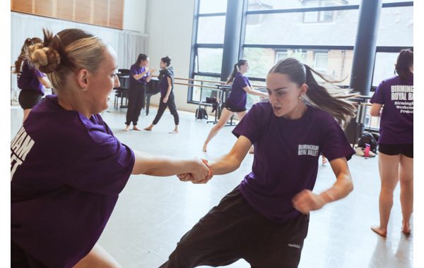 Two students are in the foreground demonstrating a jujitsu move holding hands with one trying to disentangle herself. In the background are other students trying the same move and Patsy with BRB dancer Sofia Linares talking with each other.