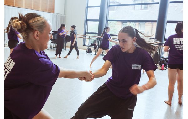 Two students are in the foreground demonstrating a jujitsu move holding hands with one trying to disentangle herself. In the background are other students trying the same move and Patsy with BRB dancer Sofia Linares talking with each other.