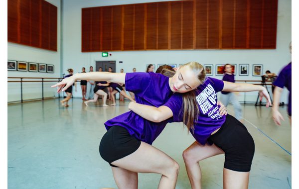 Two students are on one knee with the other leg bent leaning against each other with one arm round each other's waist and the other arm straight out in front of them. In the background are the other students.