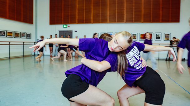 Two students are on one knee with the other leg bent leaning against each other with one arm round each other's waist and the other arm straight out in front of them. In the background are the other students.