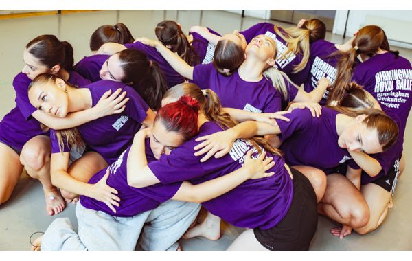 A group image with fourteen students kneeling on the floor and their arms wrapped around each other. Some are facing each others with others back to back.