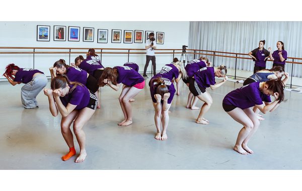 A group of students spread out in a submissive pose with both their fists in front of their faces and their legs slightly bent. Patsy and Sofia are on the right-hand side in the background observing.