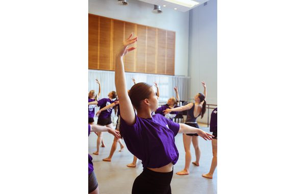 A close up of a dancer in a classical ballet pose with her left arm oustretched at ninety degrees in front of her and the other arm raised in the air with her wrist slightly bent. In the background are other students in the same pose.