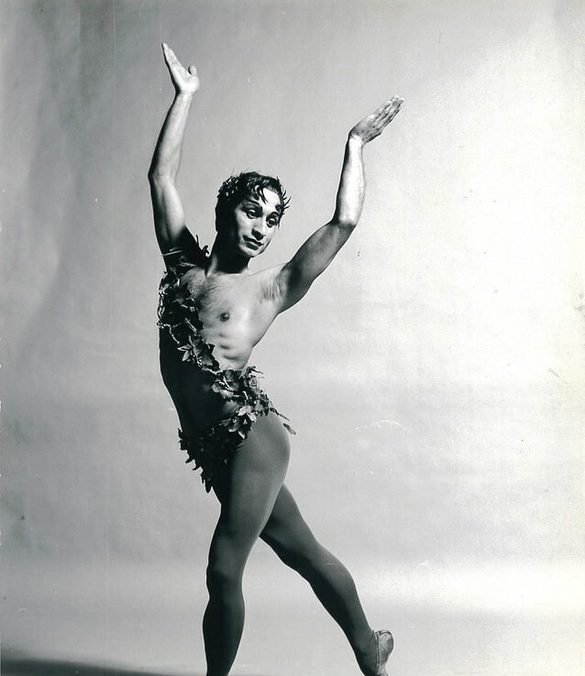 This black and white image of Johaar Mosaval pictures Johaar, the ballet legend, centre of the image with both arms raised and palms turned inwards slightly. Johaar is looking wistfully off to the right and Johaar’s dark hair which has foliage woven into it, is falling ever so slightly over the forehead at either temple. Johaar is wearing dark tights, dark ballet flats and a sash made of foliage across the chest from left hip to right shoulder. Johaar’s left leg is in front of the body and the right leg crosses behind the body, foot pointed. The background of the image is slightly textured, but blank and light in colour.