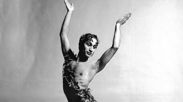 This black and white image of Johaar Mosaval pictures Johaar, the ballet legend, centre of the image with both arms raised and palms turned inwards slightly. Johaar is looking wistfully off to the right and Johaar’s dark hair which has foliage woven into it, is falling ever so slightly over the forehead at either temple. Johaar is wearing dark tights, dark ballet flats and a sash made of foliage across the chest from left hip to right shoulder. Johaar’s left leg is in front of the body and the right leg crosses behind the body, foot pointed. The background of the image is slightly textured, but blank and light in colour.