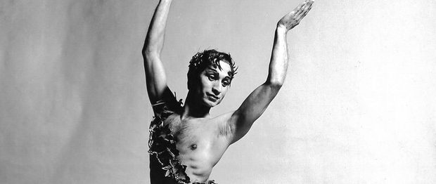 This black and white image of Johaar Mosaval pictures Johaar, the ballet legend, centre of the image with both arms raised and palms turned inwards slightly. Johaar is looking wistfully off to the right and Johaar’s dark hair which has foliage woven into it, is falling ever so slightly over the forehead at either temple. Johaar is wearing dark tights, dark ballet flats and a sash made of foliage across the chest from left hip to right shoulder. Johaar’s left leg is in front of the body and the right leg crosses behind the body, foot pointed. The background of the image is slightly textured, but blank and light in colour.