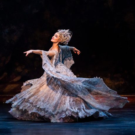 Eilis Small as the Lilac Fairy in The Sleeping Beauty. Eilis has dark hair and is wearing an opulent lilac gown made of detailed lace with a floral pattern and a headpiece made of flowers and complete with light purple jewels. Eilis is smiling and is leading with the right arm, left arm trailing behind as Eilis gracefully moves across the warmly-lit stage.