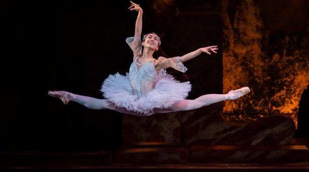 Tristram Kenton of Yu Kurihara as Princess Aurora in The Sleeping Beauty. Yu is performing a split jeté (both legs split whilst in a jump, one arm raised and one out to the side) on a stage with a golden, glowing backdrop. Yu has dark hair which is tied back and secured using an elegant tiara. Yu is wearing a light pink tutu which has sequinned details and opaque 1/4 length sleeves, pink tights and pink pointe shoes.