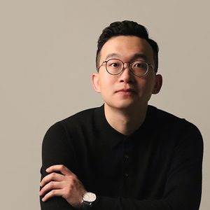 A professional headshot of Yi Wei looking at the camera against a pastel grey background. Yi Wei is sitting with arms folded, wearing a black jumper, round black glasses and a watch with a white watch face and black strap. Yi Wei has a neutral expression on their face.