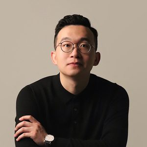 A professional headshot of Yi Wei looking at the camera against a pastel grey background. Yi Wei is sitting with arms folded, wearing a black jumper, round black glasses and a watch with a white watch face and black strap. Yi Wei has a neutral expression on their face.
