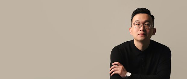 A professional headshot of Yi Wei looking at the camera against a pastel grey background. Yi Wei is sitting with arms folded, wearing a black jumper, round black glasses and a watch with a white watch face and black strap. Yi Wei has a neutral expression on their face.