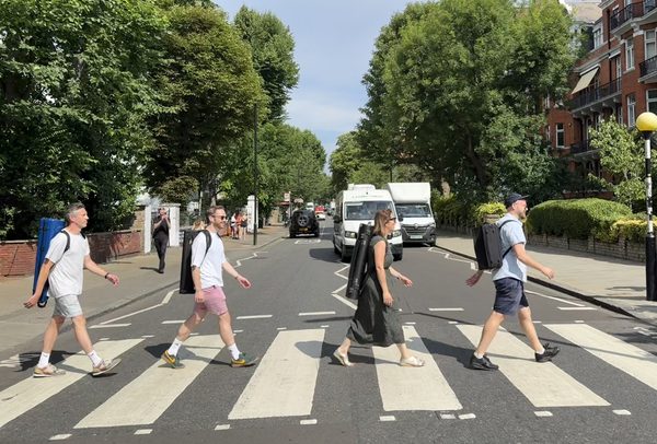 Four musicians, one in front of the other, their cased instruments on their backs, walk across a zebra crossing.