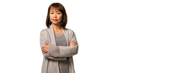 Image of BRB’s Head of Piano, Jeanette Wong, against a white background. Jeanette is standing, facing towards the camera with arms folded. Jeanette has a neutral expression. Jeanette has brown shoulder-length hair and is wearing a light brown knitted cardigan and pendant earrings.
