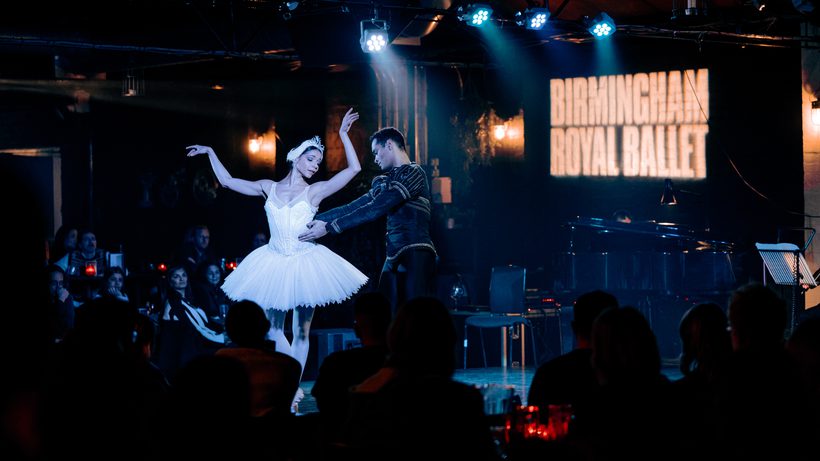 Two dancers are on a black stage at Hockley Social Club in front of a black brick wall with the Birmingham Royal Ballet logo in thick, white letters projected onto it. In front, to the left and right of the dancers are audience members seated at tables with red candles on them. The audience members in front of the dancers and to the right are stood in front of the camera, looking at the stage, so, only the backs of their heads are in the image. The audience members to the left have their faces lit by the stage lights and are also looking at the dancers. The two dancers are just off-centre of the image and are playing the role of Odette and Prince Siegfried from Swan Lake. Céline Gittens is playing Odette. Céline has black hair tied in a bun and is wearing a white feathered headband with silver tiara. Céline is wearing a white tutu, sequinned top, white tights and pale pink pointe shoes. Céline’s arms are raised, the left slightly higher than the right, Céline’s eyes are closed, with gaze pointing downwards at Siegfried. Brandon Lawrence is playing Prince Siegfried. Brandon has short black hair and is wearing a black top, decorated with gold embellishments and is also wearing black tights. Brandon is holding Céline around the waist with both arms and is facing the right of the image, gaze affixed at Céline. Above the dancers heads is a rig of blue stage lighting, which is shining down onto the dancers, creating a mystical atmosphere. Behind the dancers but in front of the wall is a music stand, and a dimly lit black baby grand piano. In the background you can see the hints of the industrial setting of Hockley Social Club, such as artificial plants and silver pipes.