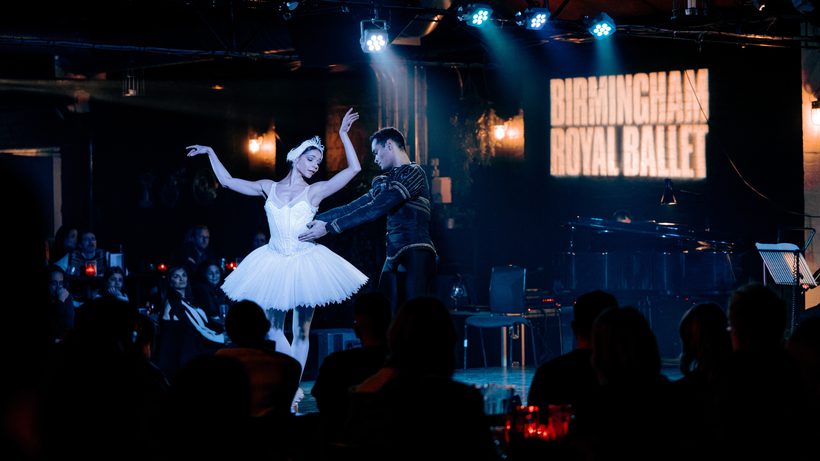 Two dancers are on a black stage at Hockley Social Club in front of a black brick wall with the Birmingham Royal Ballet logo in thick, white letters projected onto it. In front, to the left and right of the dancers are audience members seated at tables with red candles on them. The audience members in front of the dancers and to the right are stood in front of the camera, looking at the stage, so, only the backs of their heads are in the image. The audience members to the left have their faces lit by the stage lights and are also looking at the dancers. The two dancers are just off-centre of the image and are playing the role of Odette and Prince Siegfried from Swan Lake. Céline Gittens is playing Odette. Céline has black hair tied in a bun and is wearing a white feathered headband with silver tiara. Céline is wearing a white tutu, sequinned top, white tights and pale pink pointe shoes. Céline’s arms are raised, the left slightly higher than the right, Céline’s eyes are closed, with gaze pointing downwards at Siegfried. Brandon Lawrence is playing Prince Siegfried. Brandon has short black hair and is wearing a black top, decorated with gold embellishments and is also wearing black tights. Brandon is holding Céline around the waist with both arms and is facing the right of the image, gaze affixed at Céline. Above the dancers heads is a rig of blue stage lighting, which is shining down onto the dancers, creating a mystical atmosphere. Behind the dancers but in front of the wall is a music stand, and a dimly lit black baby grand piano. In the background you can see the hints of the industrial setting of Hockley Social Club, such as artificial plants and silver pipes.