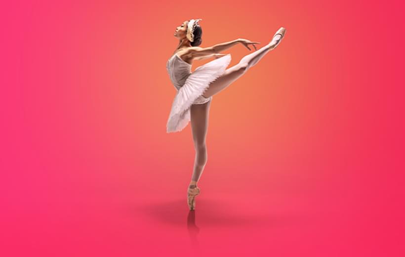Image shows a single dancer spotlighted in orange against a vivid pink background. The dancer has dark hair pulled into a bun on the back of her head. She is wearing a white tutu. The dancer is facing to the left of the image, standing on pointe on her right leg, with her right leg raised in arabesque, the toe pointing to the top right of the picture. Her arms are raised behind her in line with her raised leg.