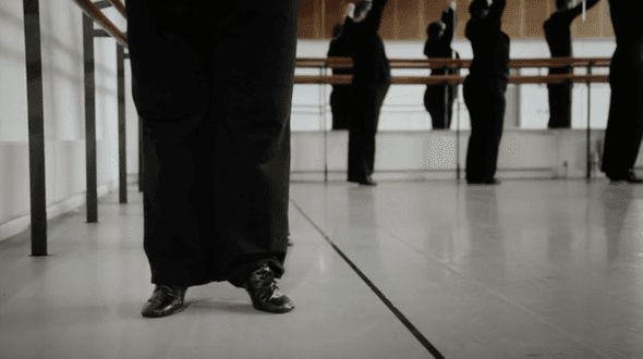Dance Studio. View is along floor. You can see four dancers, the one nearest, to the left of the image is only visible from the waist down. They are wearing black dance shows and a long black skirt or wide trousers so you can't see separate legs. The three figures further away are holding onto a ballet barre along the wall in front of a mirror. They are all holding on tiptoe with their arms above them. You can see their reflections behind them.