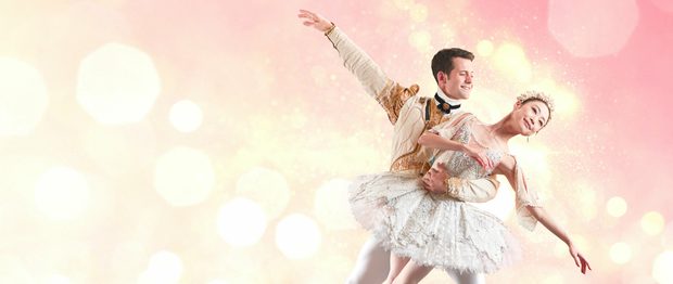 Mathias Dingman and Momoko Hirata facing towards the camera, against a pink and yellow sparkly background. Mathias is holding Momoko around the waist with the left arm. Momoko is leaning towards the right of the image, left arm draping towards the floor, right arm across the chest. Momoko is smiling and is wearing a white and tutu which has silver detailing, a decorative tiara which is holding back her dark hair, shiny earrings. Mathias is looking at Momoko smiling and right arm extended upwards. Mathias is wearing a white decorative jacket which has gold and black detailing, white tights and white ballet flats. Mathias has short dark hair. The background is a blend of blush pinks and yellow ‘sparks.’